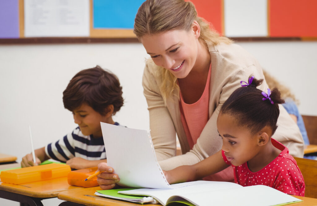 female teacher helps elementary student read in classroom